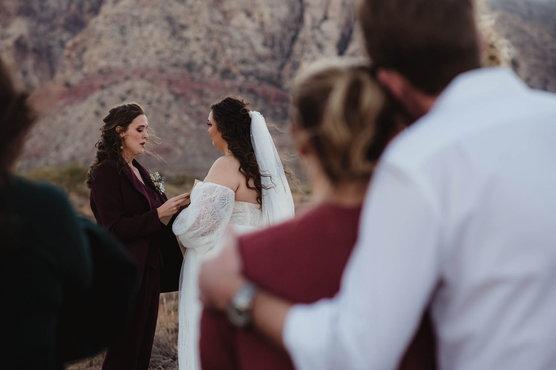 Two brunettes read vows to each other in a desert, with two people in the blurry foreground.