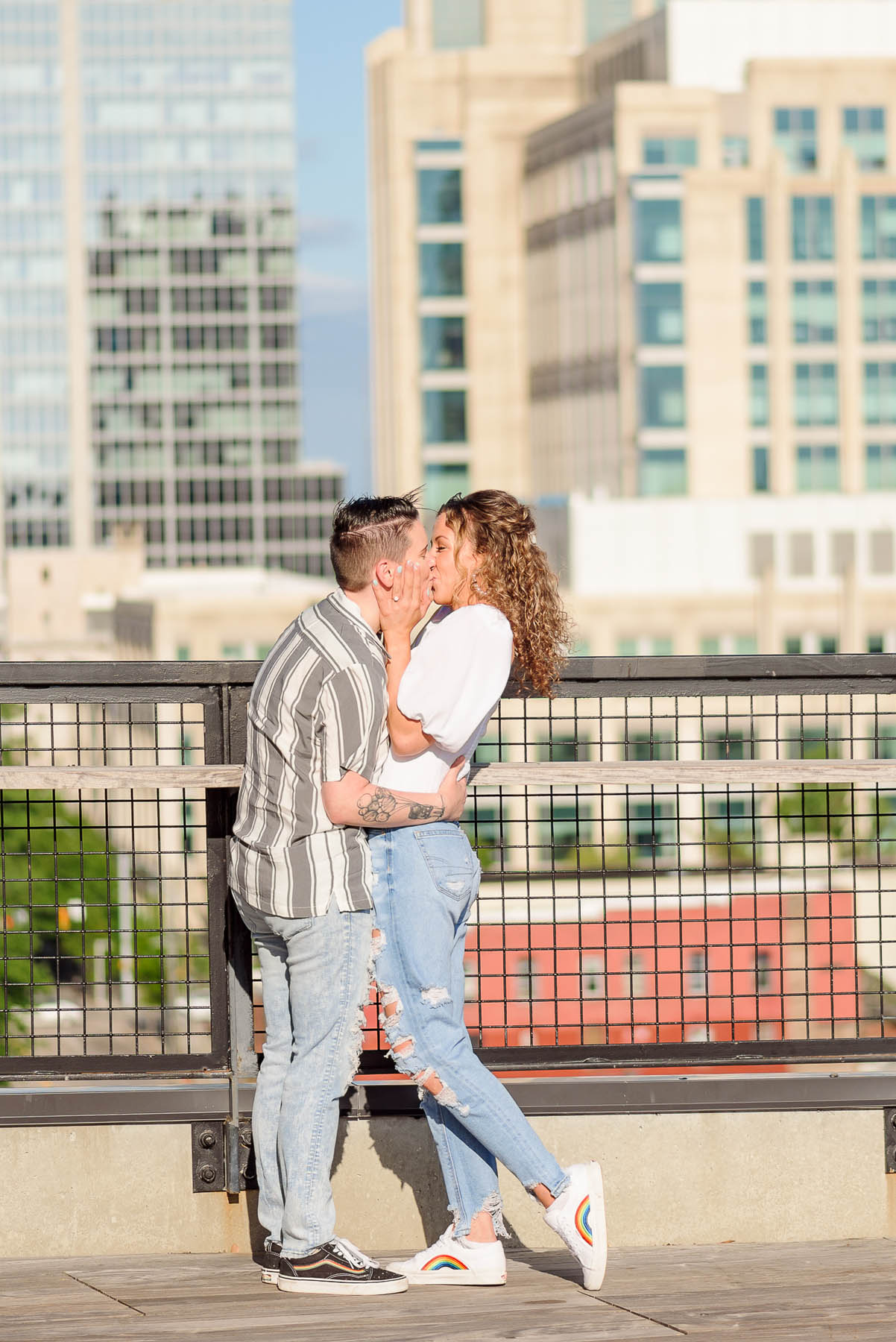 Two white people kiss in front of downtown Raleigh buildings. Both are wearing light wash jeans