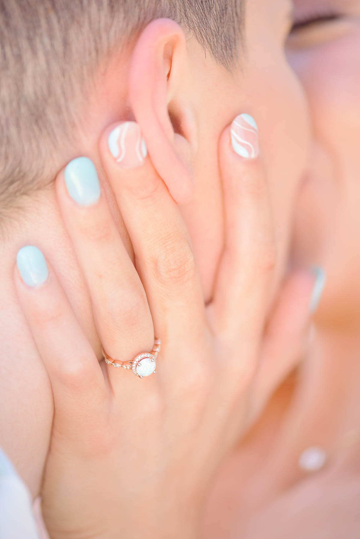 A close up oftwo white people kissing, with one's hand on the other's cheek. They are showing off a rose gold ring with an opal setting.