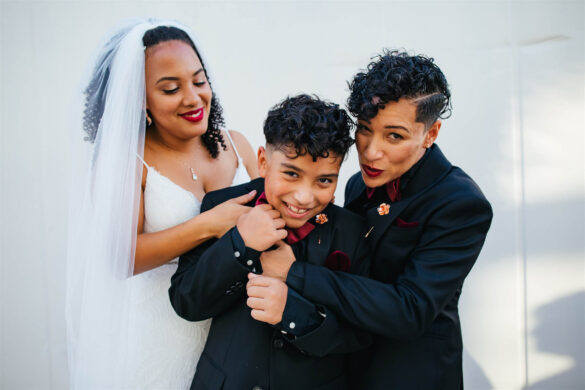 Two black brides and their son stand in front of a white wall, embracing. Two are wearing black suits and one is wearing a white gown and veil.