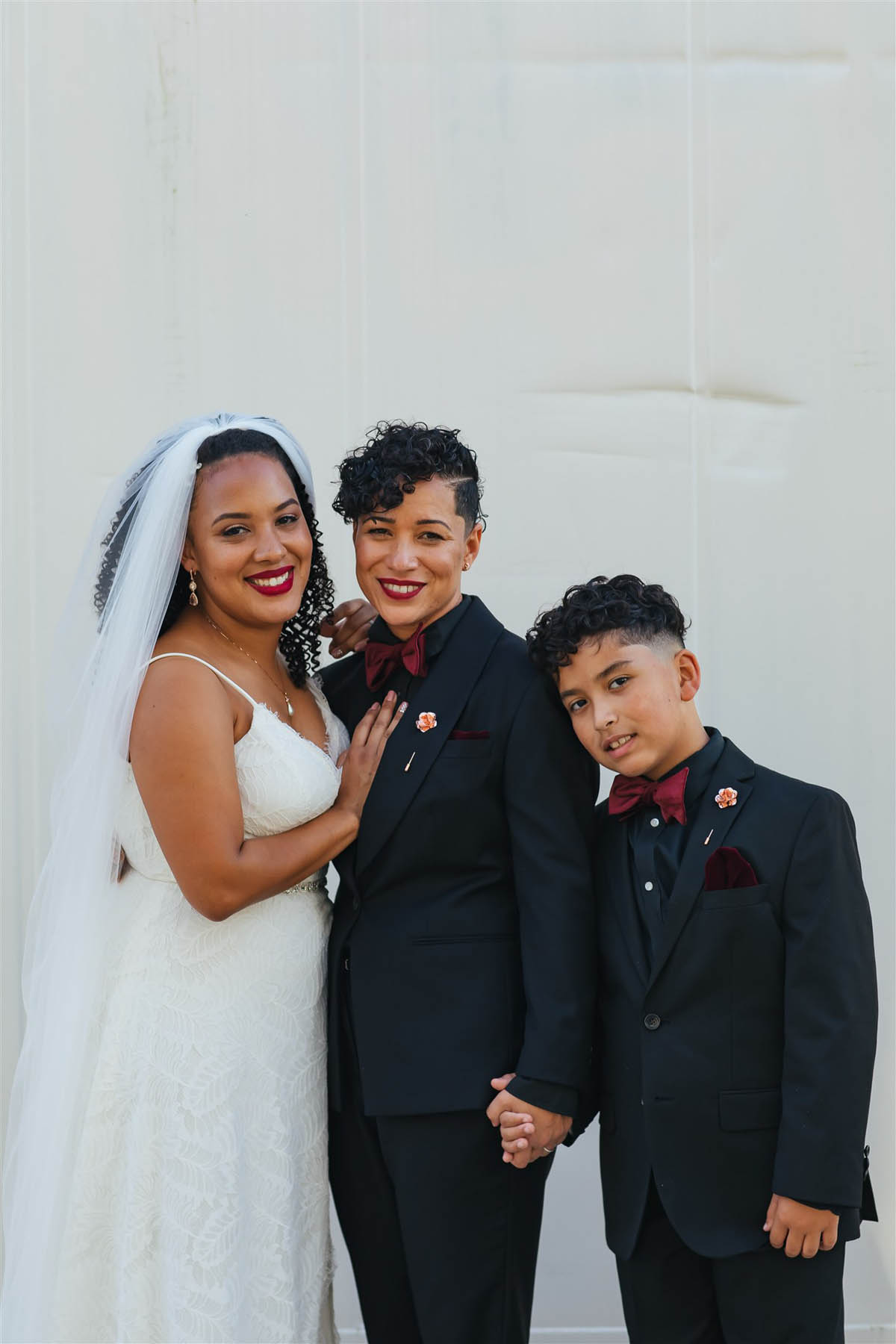 Two black brides and their son stand, holding hands, in front of a white background.