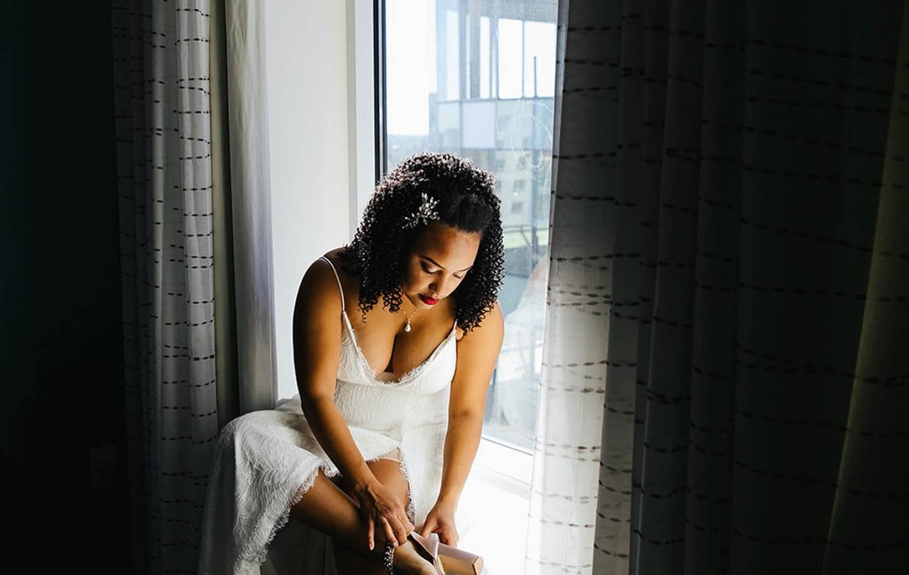 A black bride in a white gown sits on a windowsill putting on her shoes