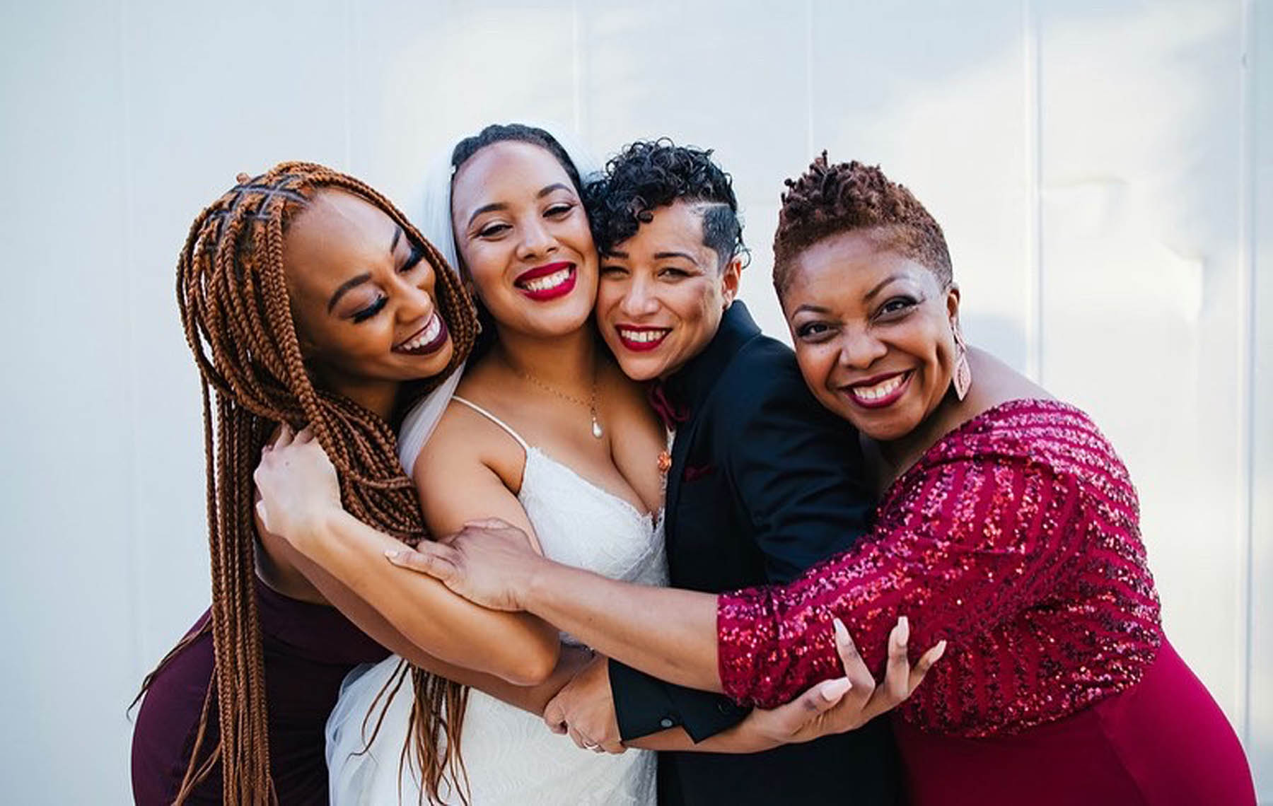 A group of four black women stand embracing in front of a white background.
