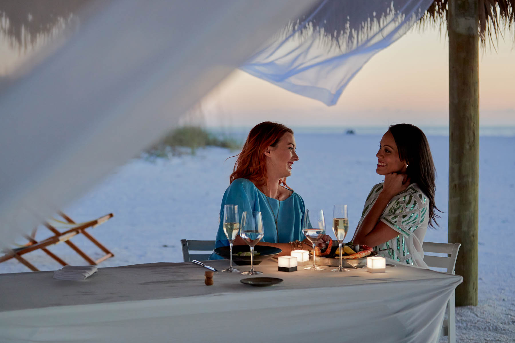 Two people with long hair sit under a beach cabana and enjoy a candlelit dinner.