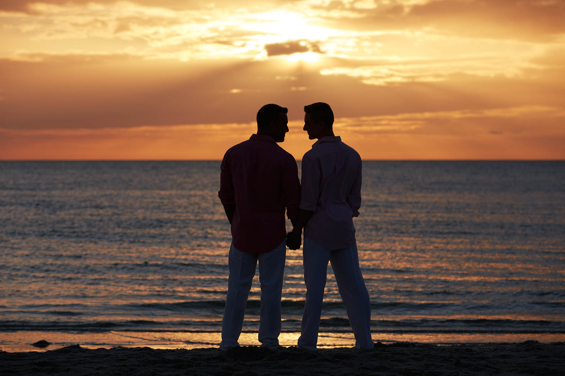 Two people stand, holding hands, against a sunset overlooking the ocean.
