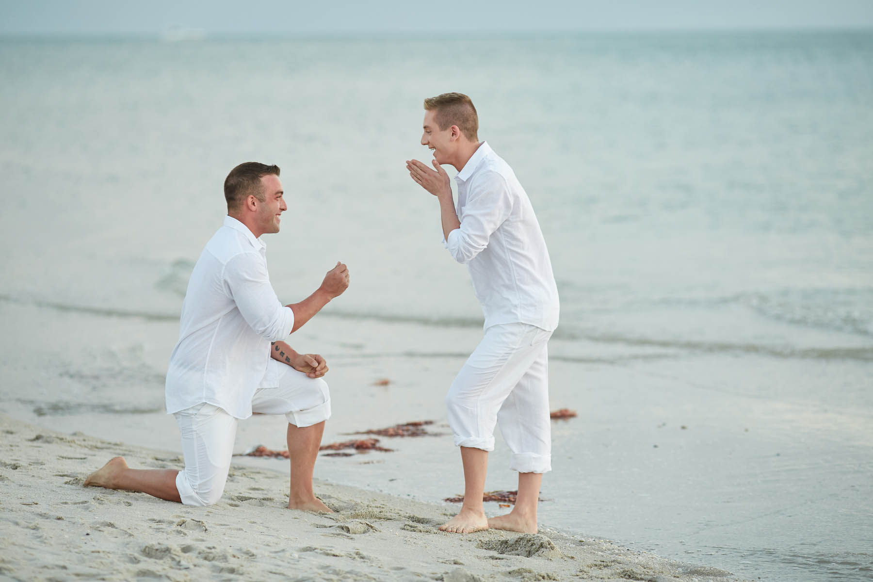 Two white people in front of the ocean, one on a bended knee proposing.