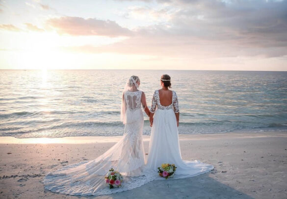 Two white people in wedding gowns stand holding hands, backs facing the camera,looking out on the ocean.