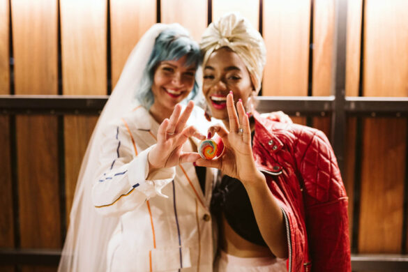 Two people, one white with short blue hair and one a person of color with a red jacket on, stand close together and hold up their hands in a heart shape for the camera.