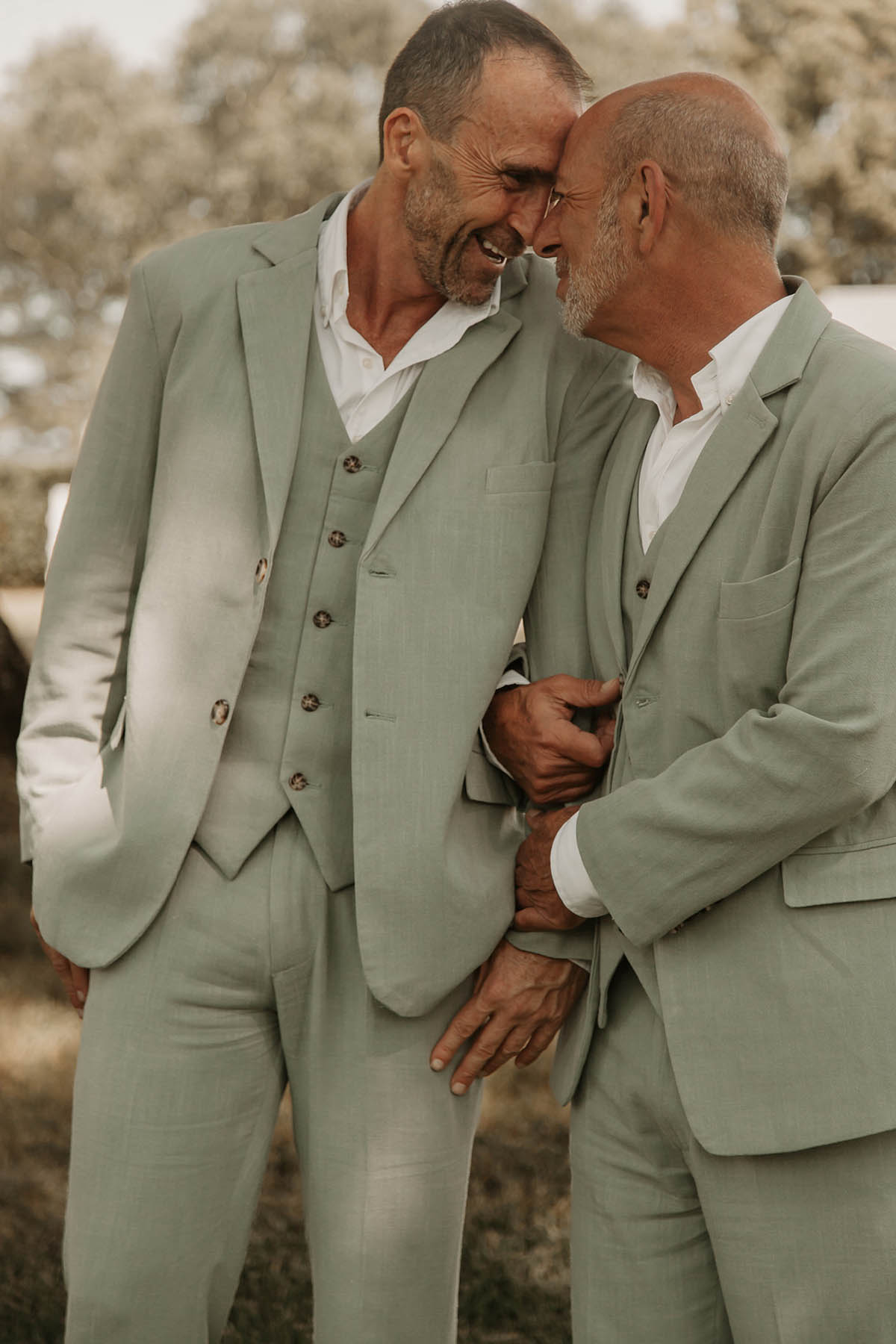 Two grooms in sage gray suits with white shirts stand arm in arm with their foreheads pressed together