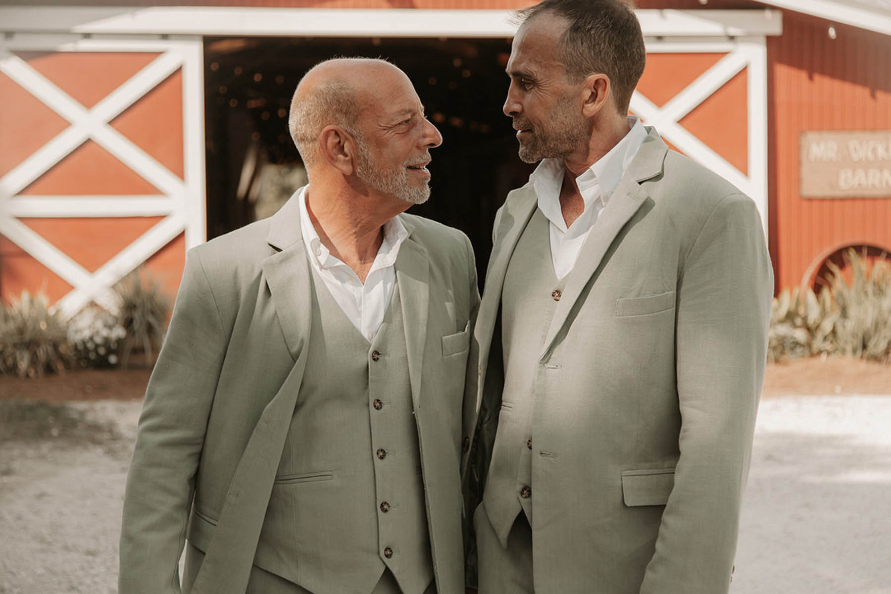 Two grooms in sage gray suits stand in front of an open red barn, smiling at each other