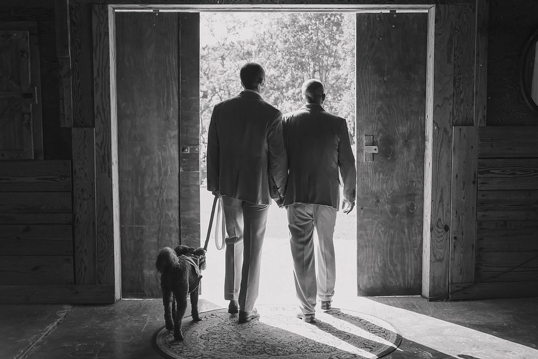A black-and-white photo of two grooms walking with their dog alongside them.