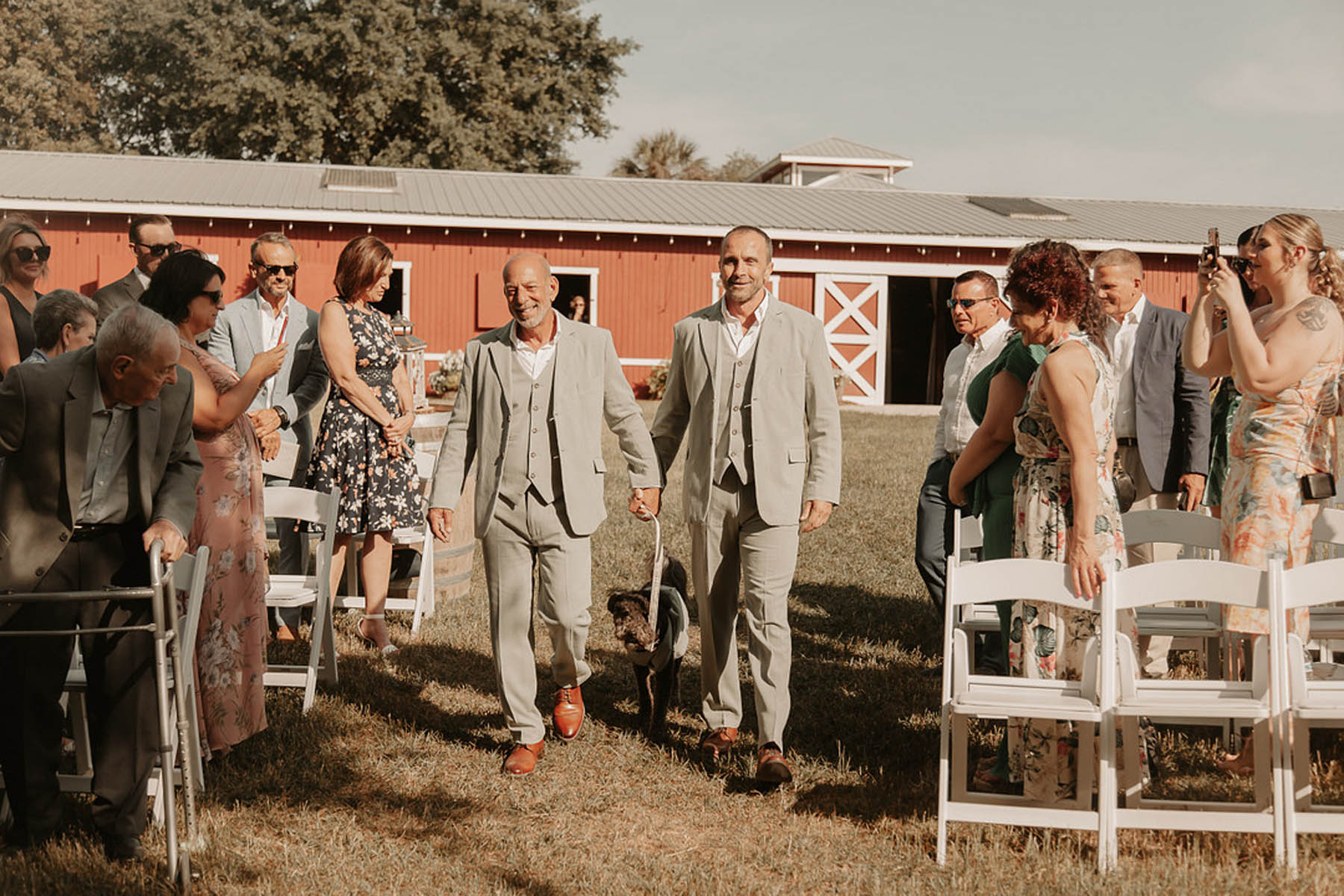 Two grooms in sage gray suits walk with their dog down an aisle in front of a red barn