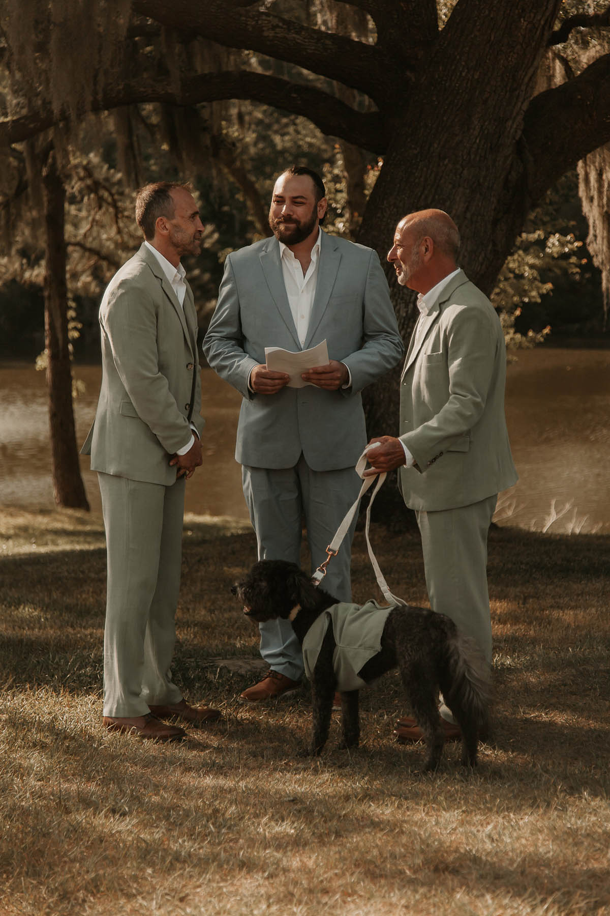 Two grooms in sage gray suits stand in front of a large tree with their officiant, a white person in a blue suit.