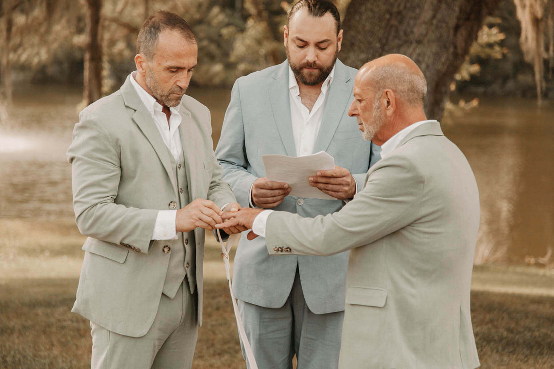 Two grooms in sage gray suits and exchange rings in front of their officiant, a person in a blue suit.