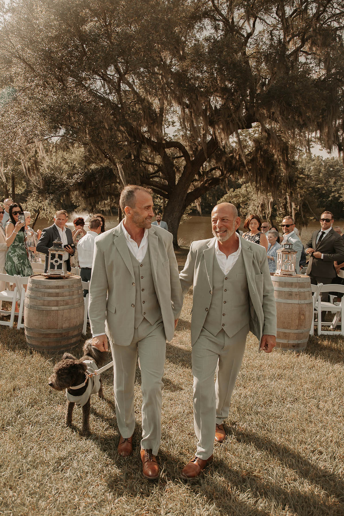 Two grooms in sage gray suits walk hand in hand, with their dog by their side, down an aisle with a large tree in the background.