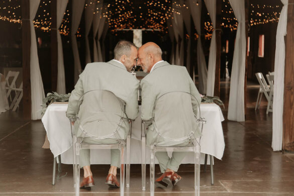Two white grooms in sage gray suits sit at a table in clear chairs with their foreheads touching.