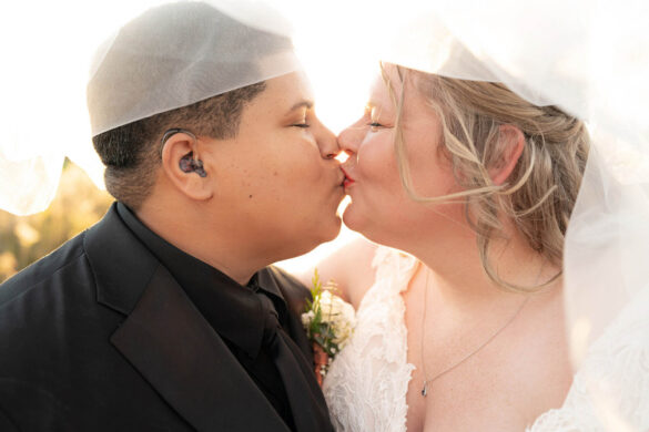 Two brides, one white with long blonde hair and one black with short dark hair, share a kiss under a white veil.