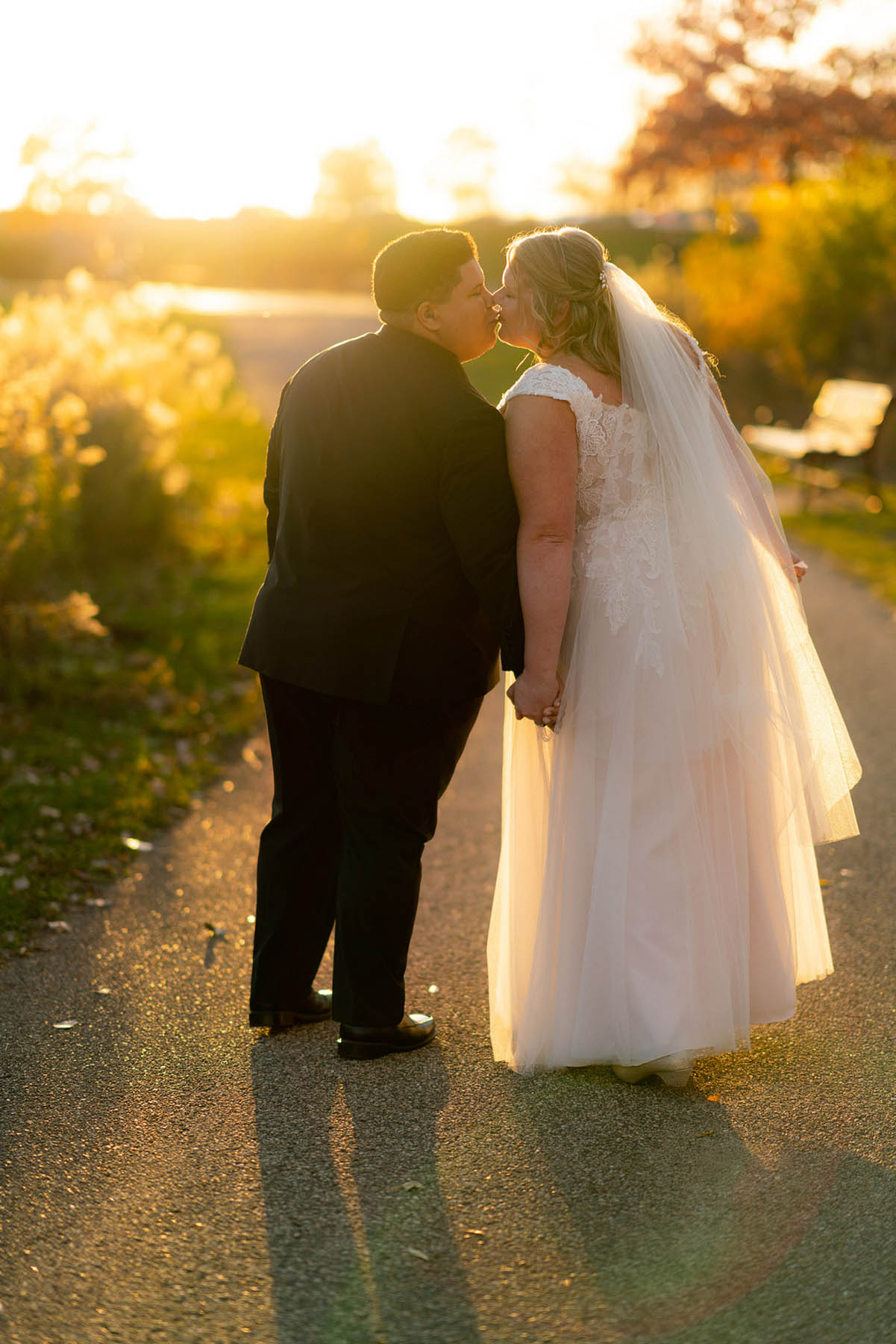 Two brides, one white in a white gown and one black in a black suit, share a kiss on an outdoor path with fall foliage in the background.