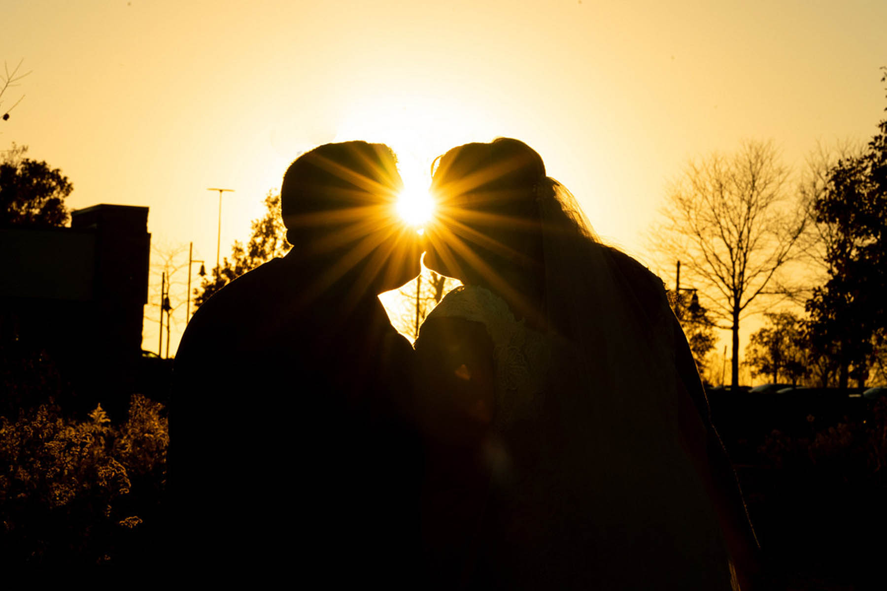 Two figures share a kiss with the setting sun between then, creating a lens flar at the center point between them.