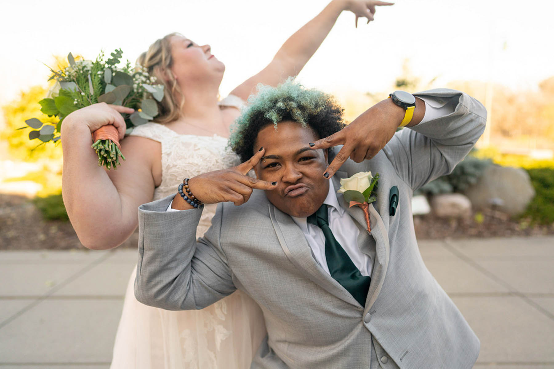 A white woman in a white gown stands behind a black person in a gray suit with green hair, striking a funny and expressive pose. 