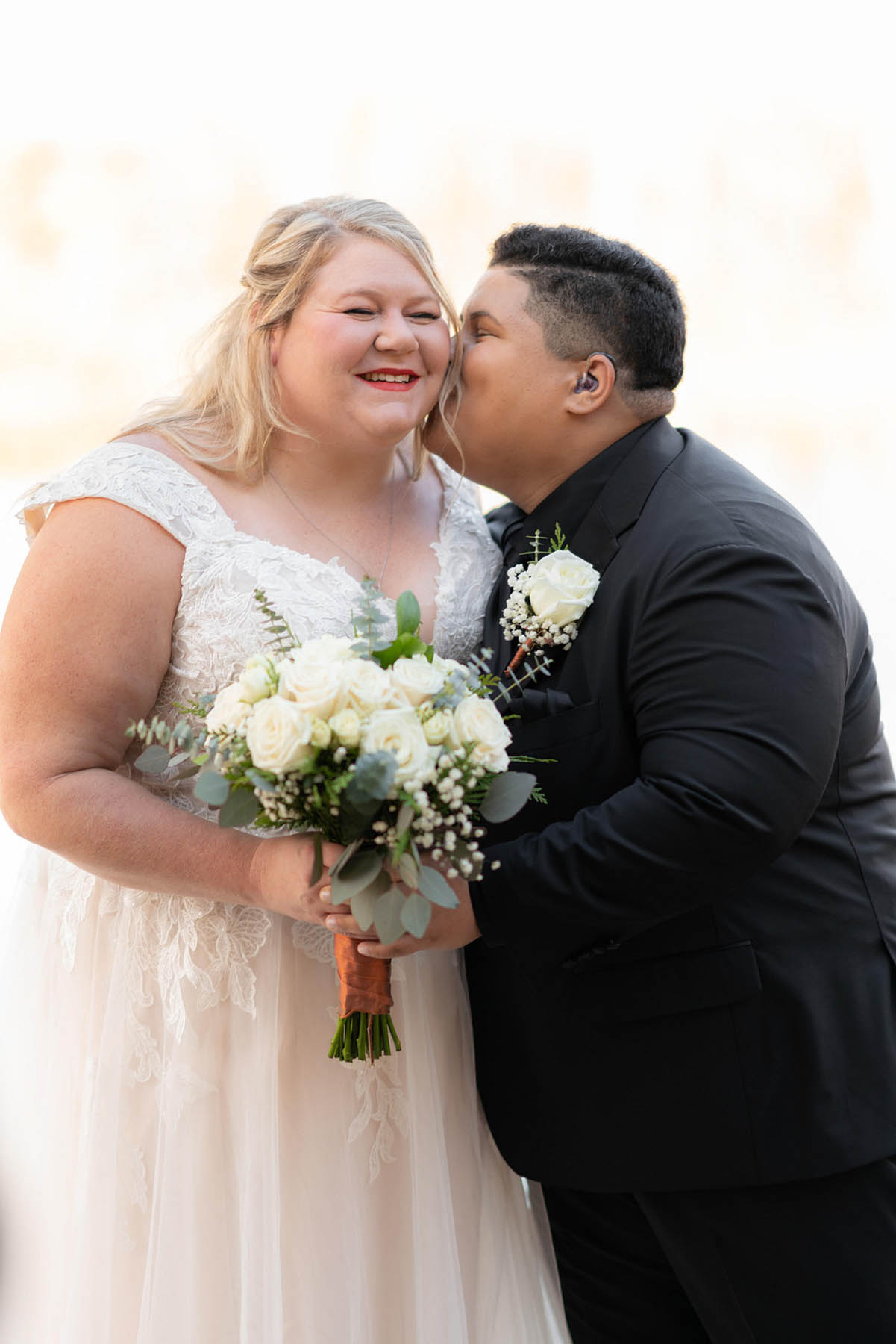 Two brides, one white and one black, stand arm in arm next to each other, as one kisses the other on the cheek