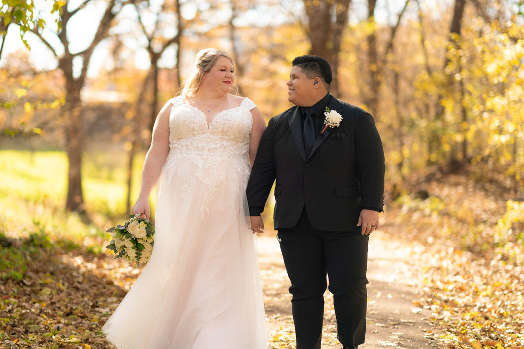 Two brides, one white in a white gown and one black in a black suit, walk hand in hand down an outdoor path with fall foliage in the background.