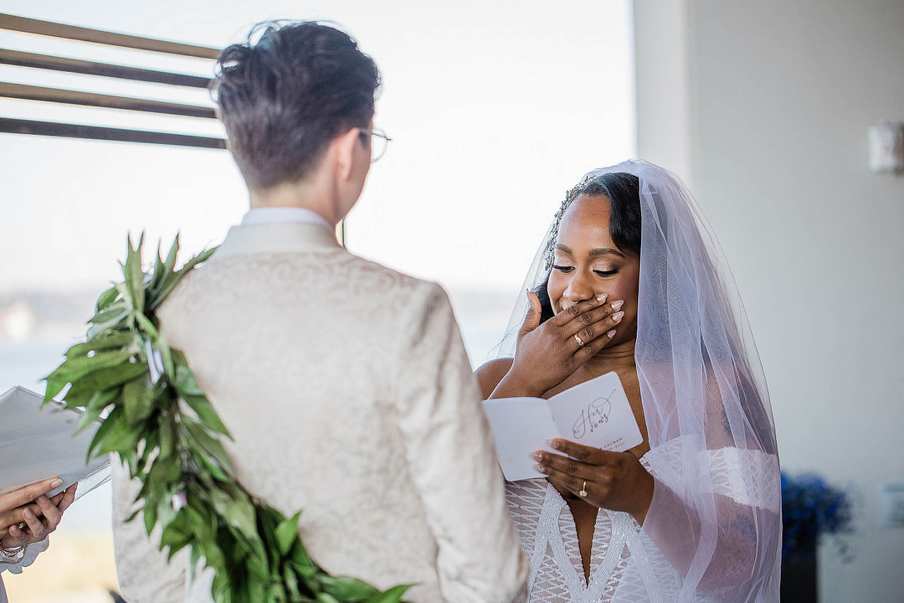 Two brides, one black and one white, stand facing each other in front of a large window, reading vows.