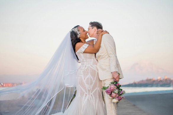 Two brides, one white and one black, stand in front of a sunset and share a kiss.
