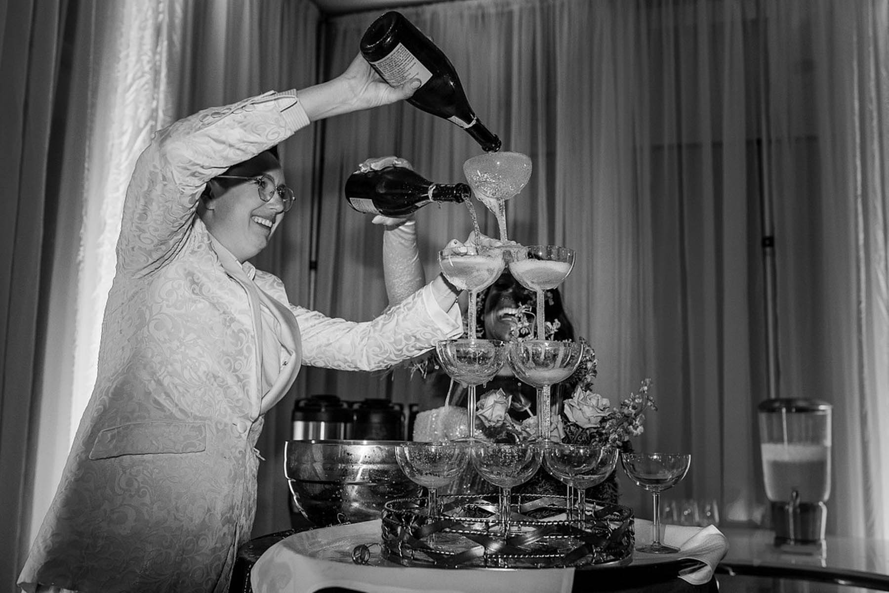 A black-and-white photo of the marriers pouring champagne on a champagne coupe tower.