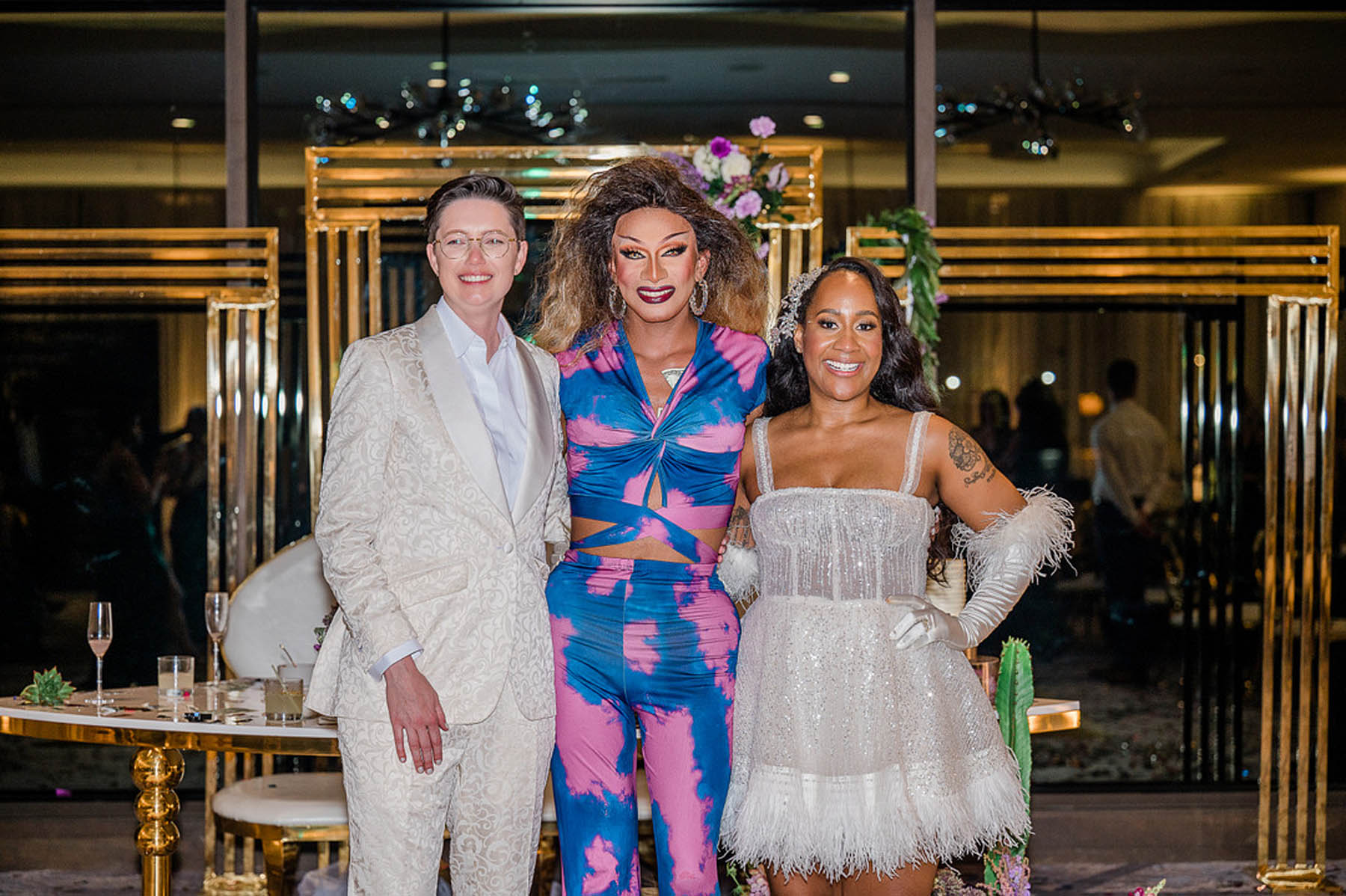 The marriers stand on either side of a drag queen, who is dressed in a blue and pink jumpsuit.