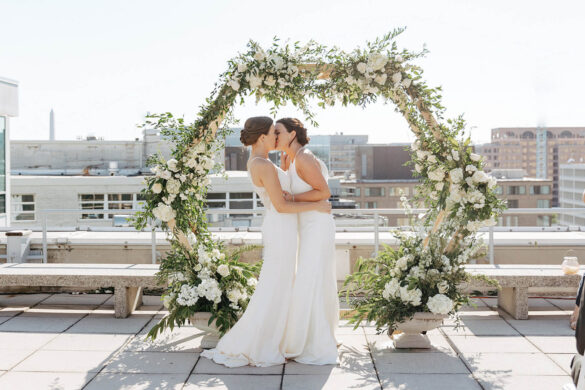 Two white brides in long white dresses share a kiss at their rooftop ceremony.
