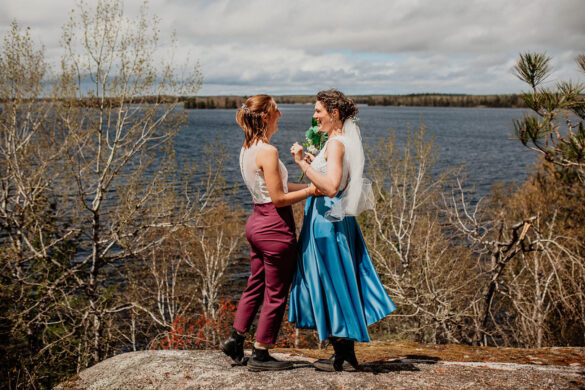 Two white brides stand in front of a lake and exchange vows