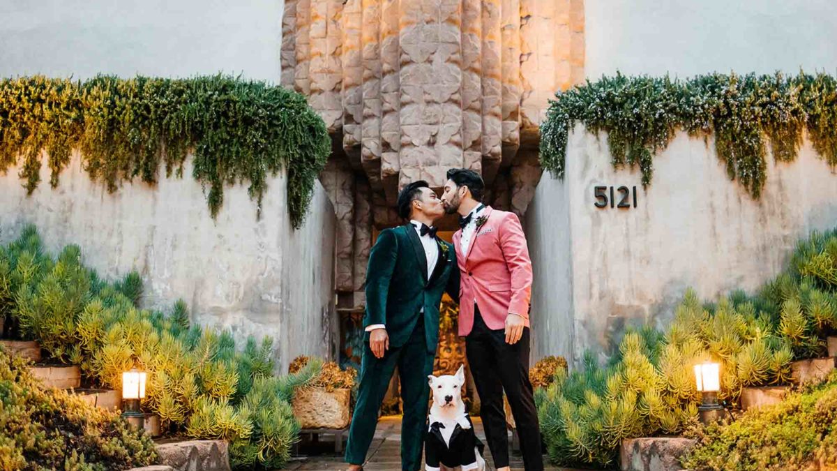 Two grooms, in green and pink jackets, kiss with their dog in between them.