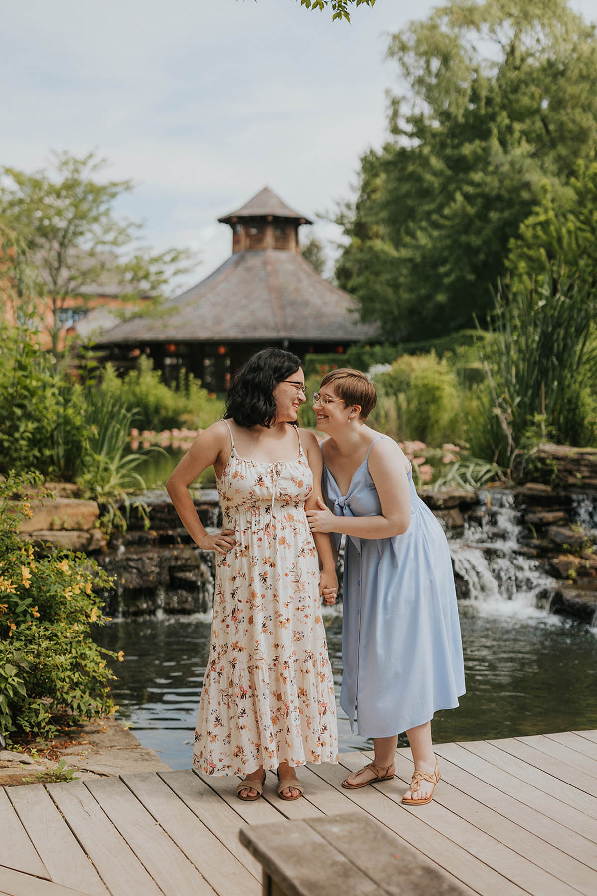 Two femme people stand in front of a pond and gazebo. They are laughing and smiling at each other.