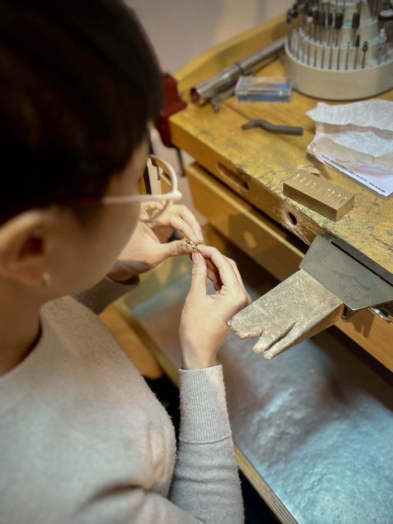 A person with glasses on sits at a workbench and crafts metal jewelry.
