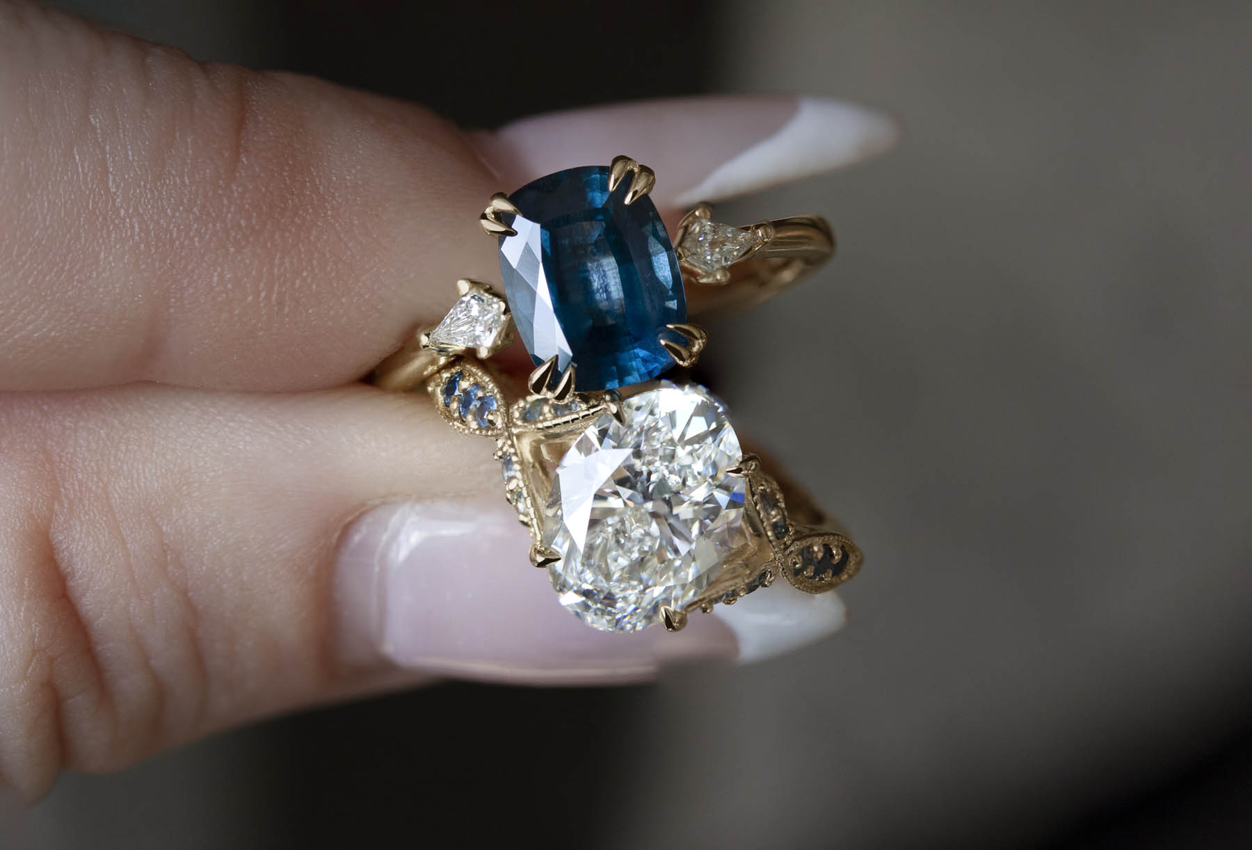 A close-up of two rings: one with a blue rectangular cut gem, and one with an oval, crystalline gem. 