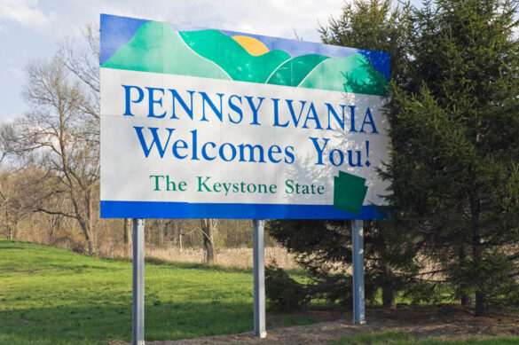 A sign with green and blue accents in front of a green patch of grass and trees, reading: Pennsylvania welcomes you! The Keystone State