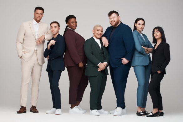 7 people wearing suits. From left to right, the suit colors are light tan, navy, burgundy, forest green, navy, blue and black. The models are a variety of races and ethnicities, gender identities and sizes.