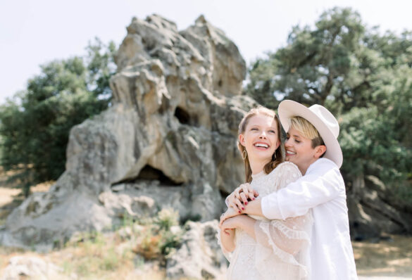 Two white marriers hug in front of a large boulder. One wears a white dress with gold earrings, and the other has a white wide brim hat.