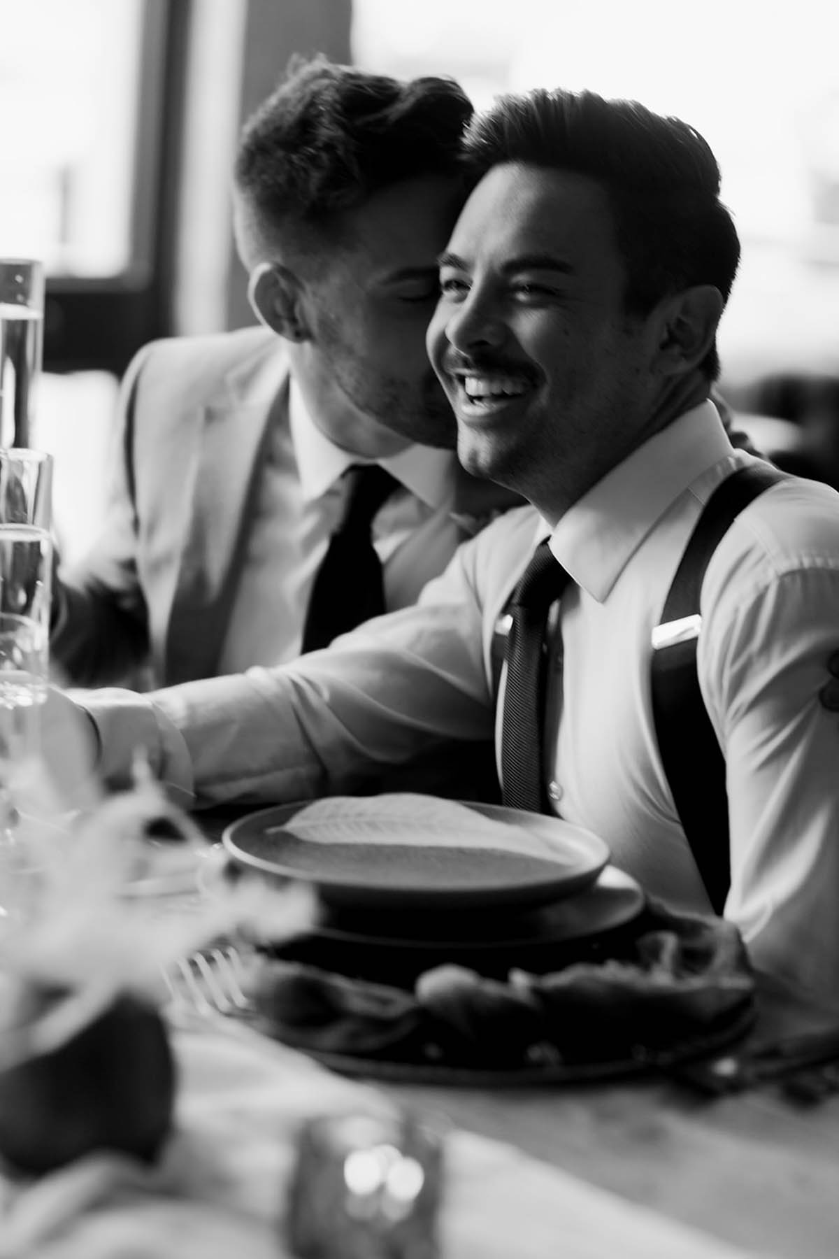 A black-and-white photo of two grooms sitting at their reception table. One is talking in the other's ear. The other is smiling.