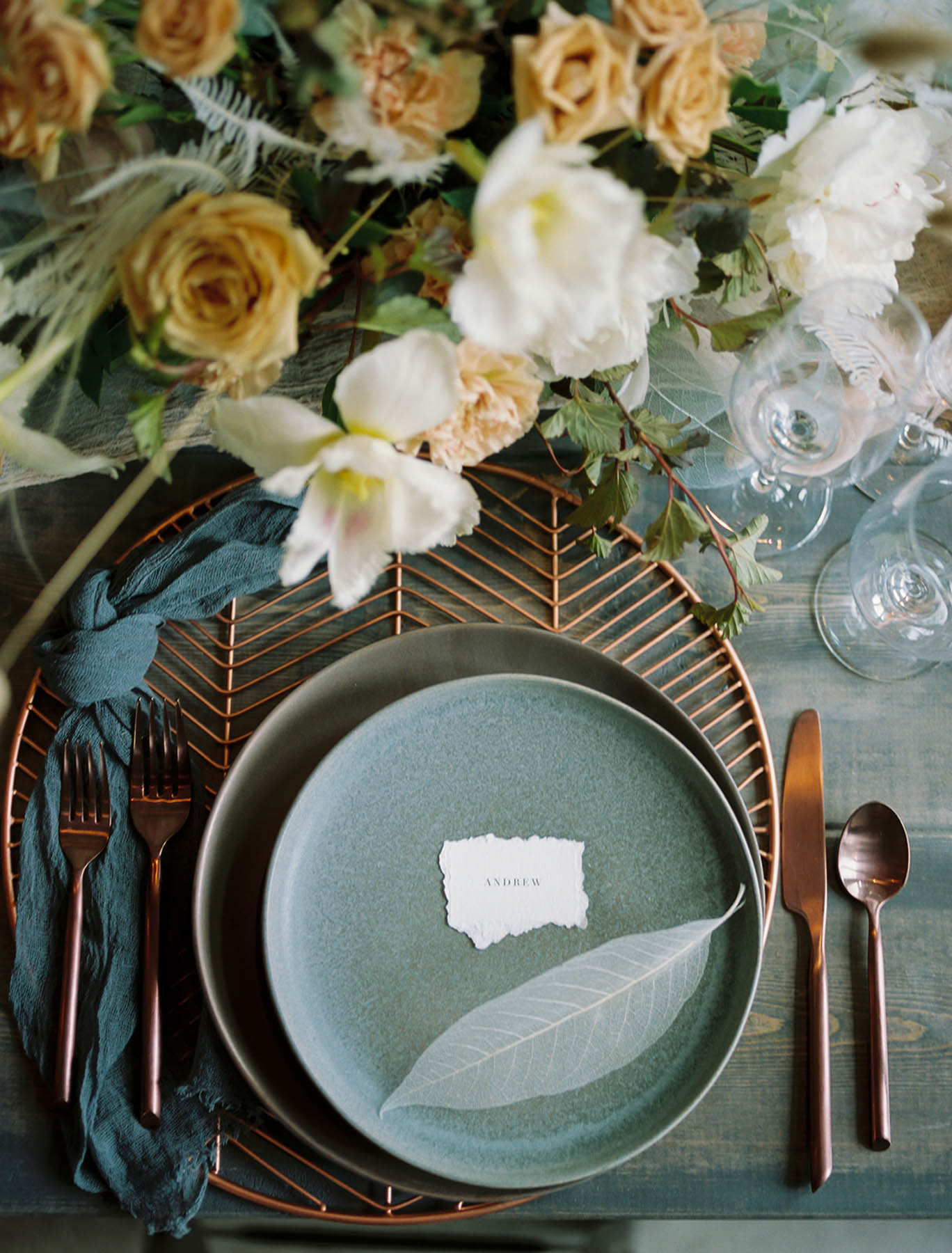 A place setting with white florals and gray-green elements. There is bronze silverware and a small place card on the center of the plate. 
