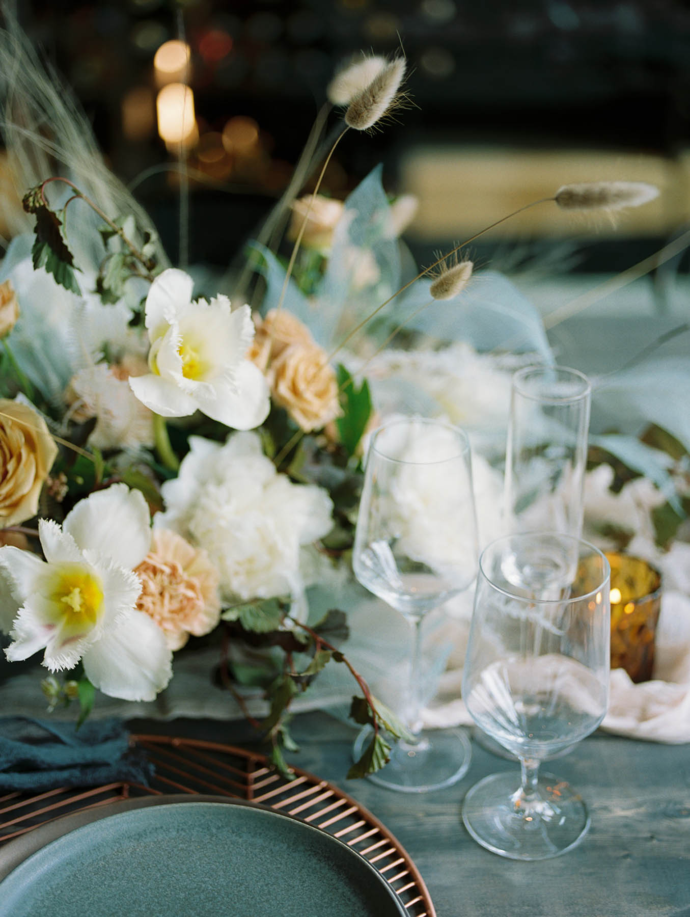 A close-up on a tablescape of white and pink flroals and blue cloth. There is a gold candle and clear glasses.
