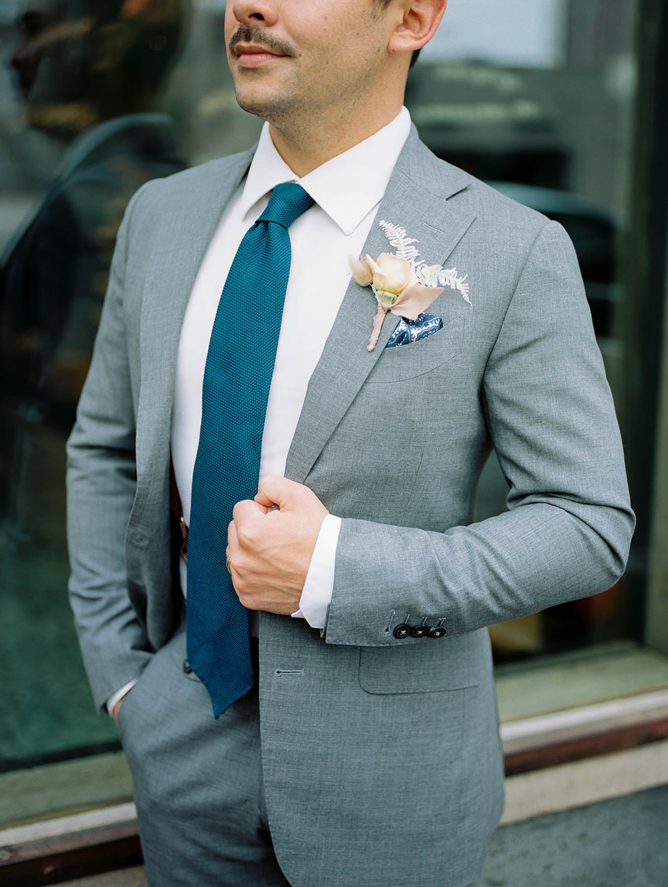 A white groom with a blue tie stands with one hand on his lapel and one in his pocket. 