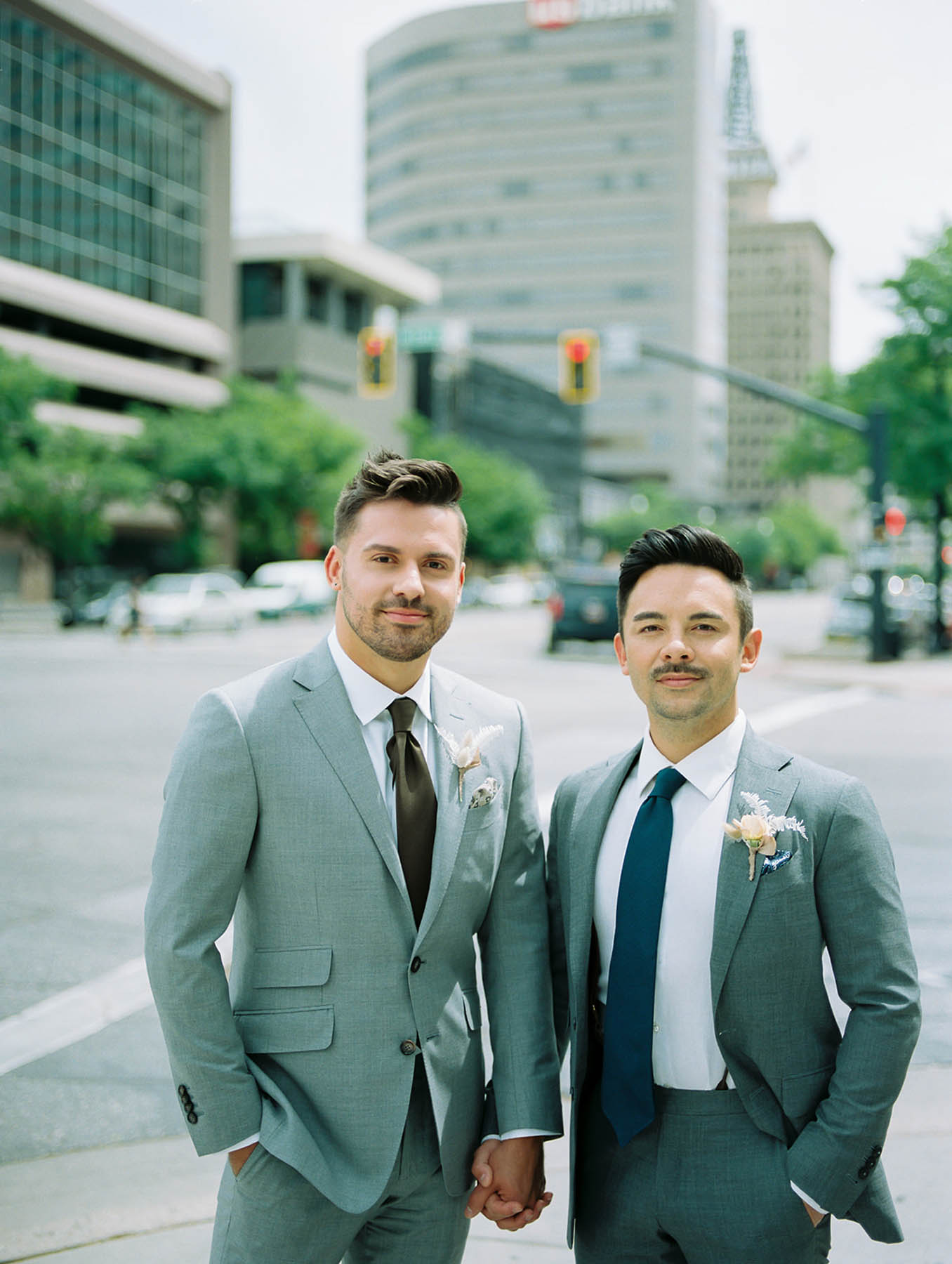 Two white grooms with dark hair and gray suits stand on a city street in Park City. 
