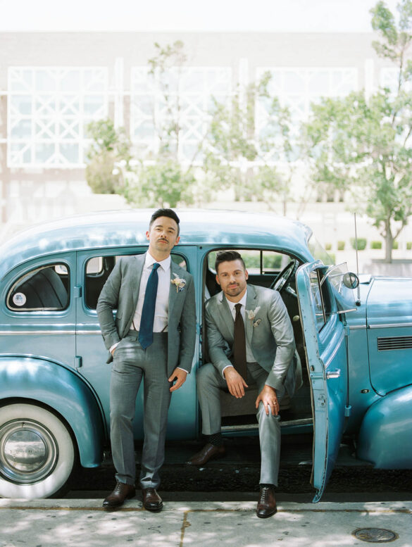 Two white grooms in gray suits are in front of a vintage blue car. One sits in the passenger seat with the door open, and the other stands, leaning against the car.