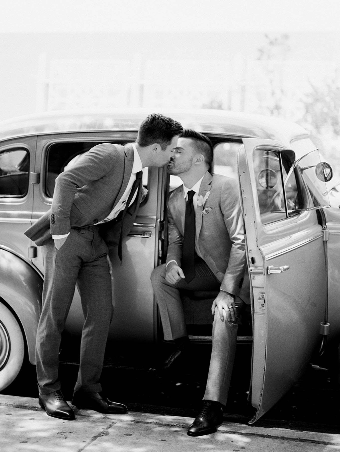 A black-and-white photo of two white grooms kissing next to a vintage car.