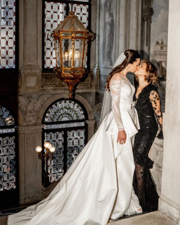 Two white brides kissing. One woman is wearing a long-sleeved white wedding gown, and the other woman is wearing a long-sleeved black wedding gown.