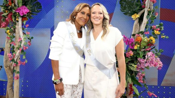 Celebrity lesbian couple Robin Roberts, a Black woman with honey-colored hair, is wearing a white jacket and a bride-to-be silver sash, and Amber Laign, a white woman with blonde hair who is wearing a white outfit and an identical silver sash. Bright flower displays are on either side of them.