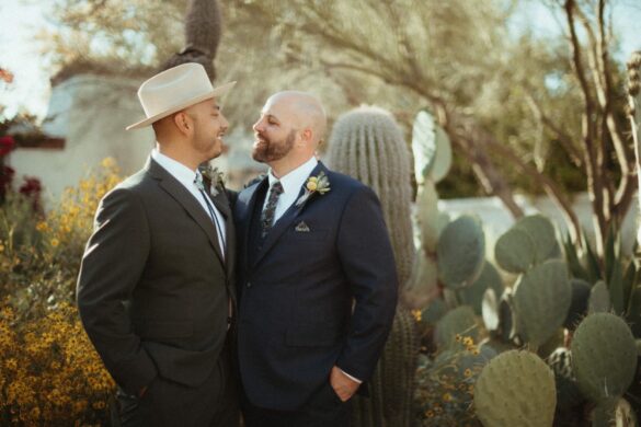 Two grooms smile at each other. They're wearing dark suits and standing in front of a cactus.