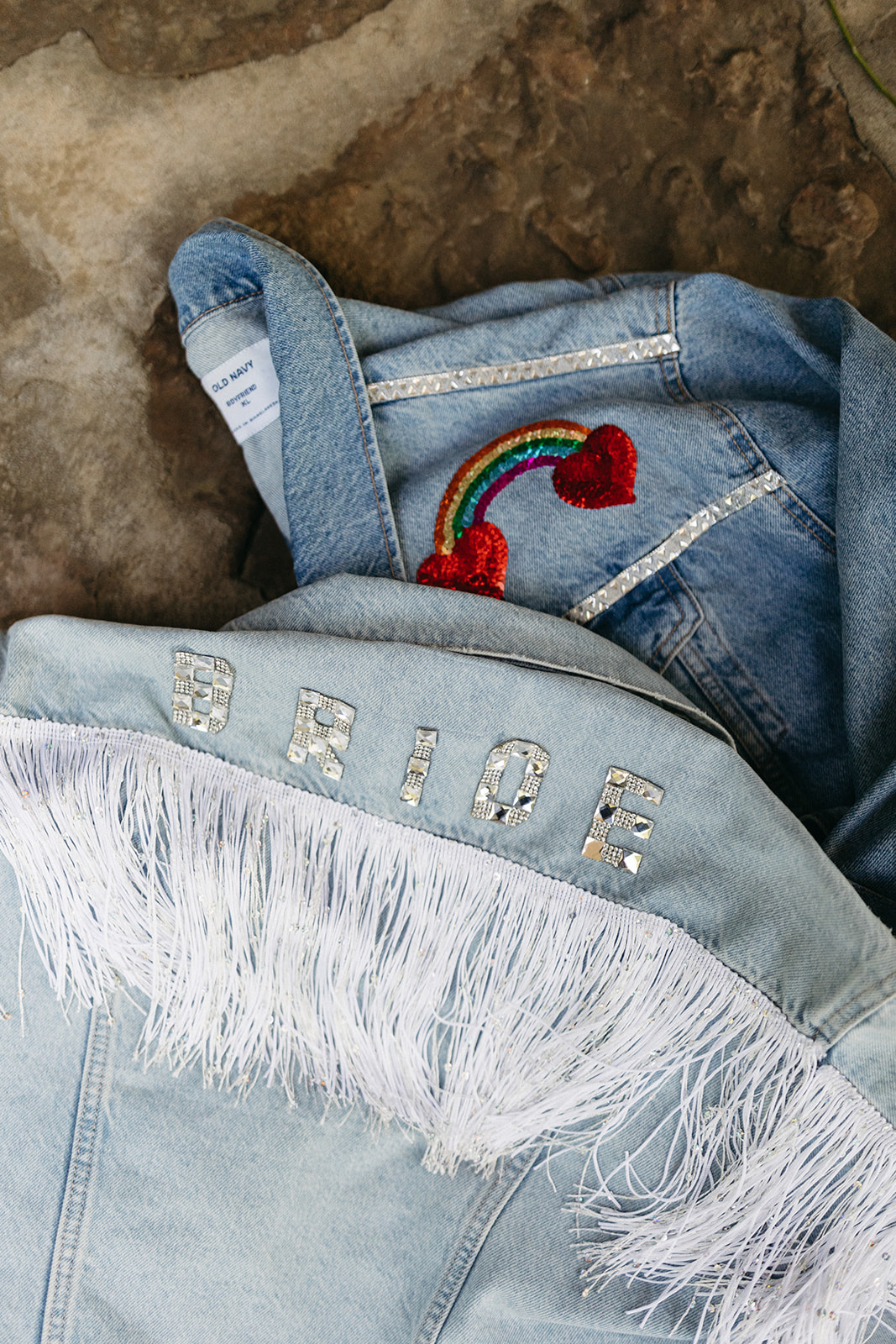 Denim jackets for two brides. One has silver letters spelling out bride with white fringe underneath. The other jacket has a sequin rainbow and two red hearts.